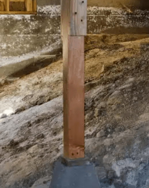 A wooden post stands upright on a small concrete base in a basement with unfinished walls and a dirt floor. The dim lighting highlights the rough texture, emphasizing the need for basement waterproofing solutions in Bergen County, NJ.