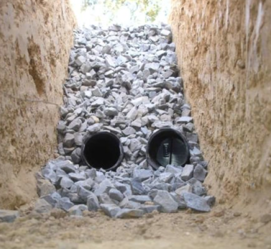 A drainage system under construction, featuring two pipes partially embedded in a trench. This vital part of basement waterproofing services in Bergen County, NJ, includes pipes surrounded by crushed stone, with trench walls of compacted earth. Sunlight illuminates the background.