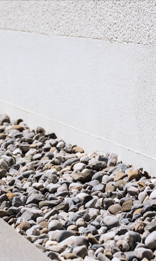 A strip of small, rounded gray and brown pebbles lies alongside the base of a white stucco wall. The sunlight casts soft shadows, highlighting the texture of the rocks and the wall—much like how experts in basement waterproofing in Bergen County NJ protect homes from unseen challenges.