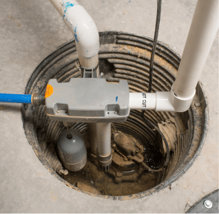 A sump pump system is installed in a basement floor for effective water drainage. The image shows pipes and a pump partly submerged in the pit, ensuring basement waterproofing in Bergen County, NJ. The concrete surroundings have some visible water at the bottom of the pit.