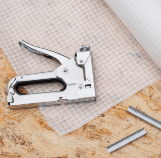 A silver staple gun rests on a wooden surface beside a roll of white mesh material, with metallic staples scattered nearby. The setup hints at preparation for a DIY project or fabric installation, perhaps part of basement waterproofing in Bergen County NJ.
