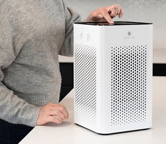 A person in a gray sweater operates a white air purifier on a countertop, preparing for basement waterproofing in Bergen County NJ. The purifier flaunts a perforated design and a logo on top. The person's right hand is on the device, while the left hand rests casually on the counter.